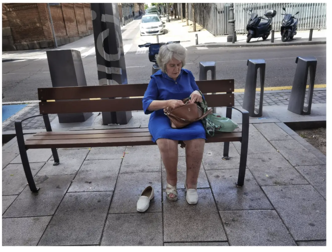 elderly woman sitting on public seat with one shoe off, in Madrid Spain