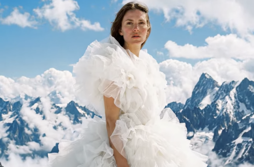 AI-generated portrait created with Google Veo 3 showing a woman in a dramatic white ruffled dress standing on a snow-covered mountain ridge beneath a vivid blue sky with towering alpine peaks and clouds in the background.