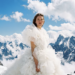 AI-generated portrait created with Google Veo 3 showing a woman in a dramatic white ruffled dress standing on a snow-covered mountain ridge beneath a vivid blue sky with towering alpine peaks and clouds in the background.