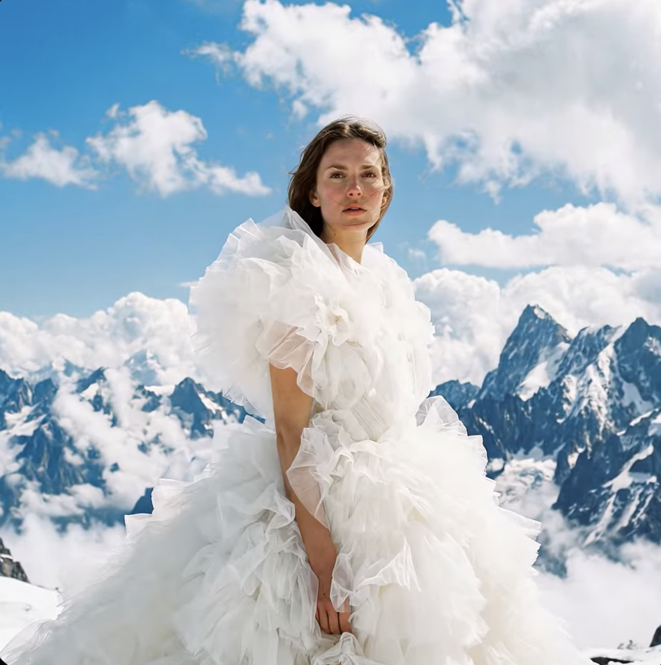 AI-generated portrait created with Google Veo 3 showing a woman in a dramatic white ruffled dress standing on a snow-covered mountain ridge beneath a vivid blue sky with towering alpine peaks and clouds in the background.