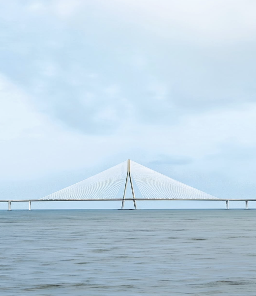 Minimalist seascape featuring a perfectly centred cable-stayed bridge stretching across calm blue water beneath a soft, cloud-filled sky. The bridge's symmetrical white cables form a striking geometric composition, creating a serene, almost abstract architectural landscape.