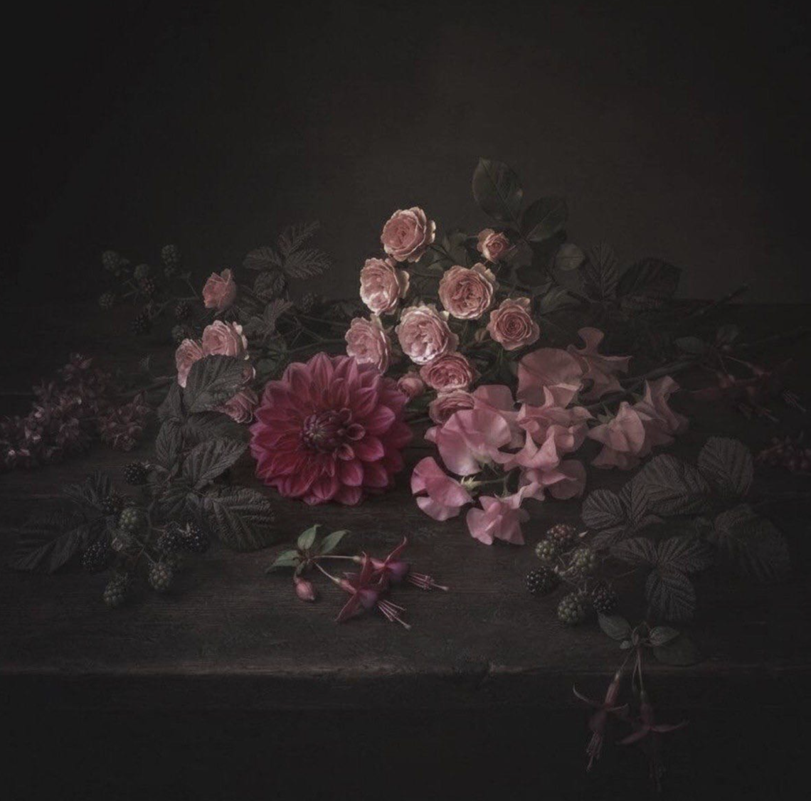 A low-key still life featuring a dramatic arrangement of pink roses, a deep magenta dahlia, delicate sweet peas, blackberries, fuchsia blossoms and dark foliage resting on a rustic wooden table. Soft directional light illuminates the flowers against a rich black background, creating a timeless, Dutch Master-inspired composition with a moody, painterly atmosphere.