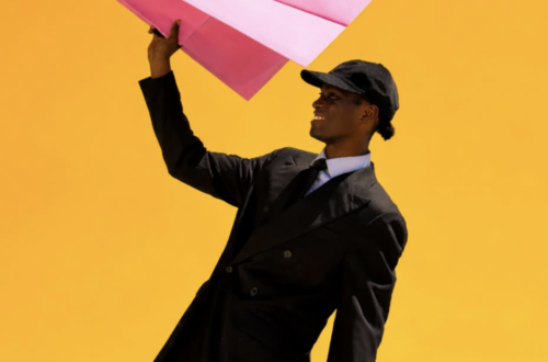 A fashion portrait of a smiling person in an oversized black suit, tie and cap holding a large pink paper aeroplane aloft against a vivid golden-yellow background, creating a playful, graphic composition with bold colour contrast.