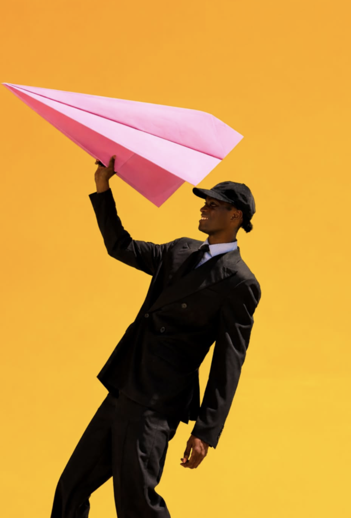 A fashion portrait of a smiling person in an oversized black suit, tie and cap holding a large pink paper aeroplane aloft against a vivid golden-yellow background, creating a playful, graphic composition with bold colour contrast.