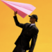 A fashion portrait of a smiling person in an oversized black suit, tie and cap holding a large pink paper aeroplane aloft against a vivid golden-yellow background, creating a playful, graphic composition with bold colour contrast.