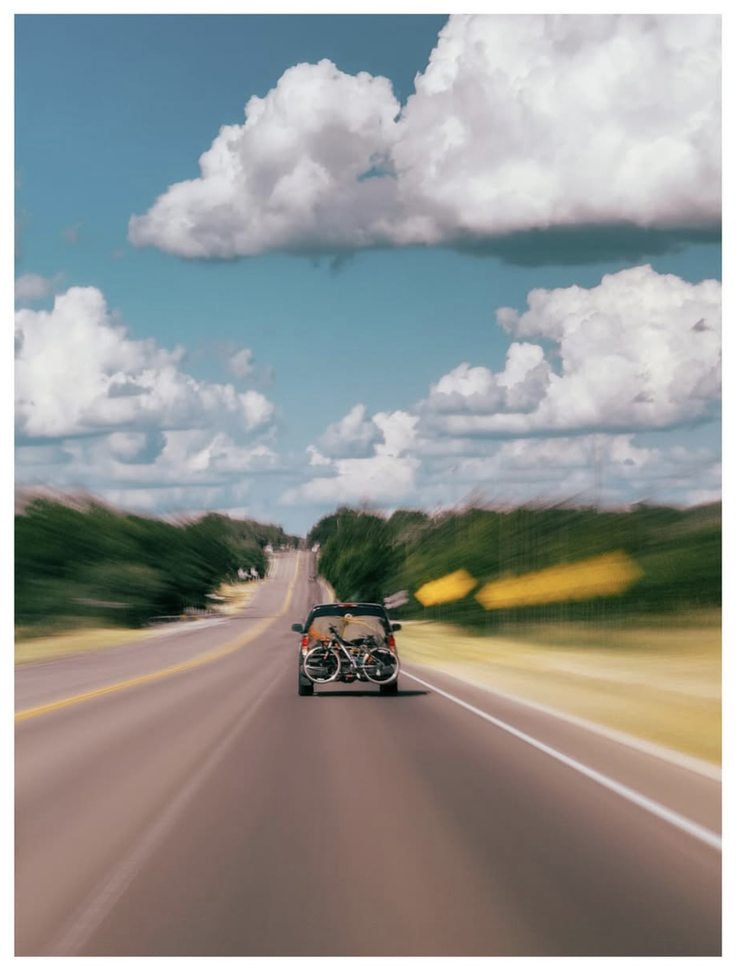 A motion-blurred photograph of a car carrying two bicycles on a rear rack driving along a winding rural highway beneath towering white cumulus clouds. The dynamic zoom effect emphasises speed while the expansive blue sky and rolling green landscape create a sense of adventure and open-road travel.