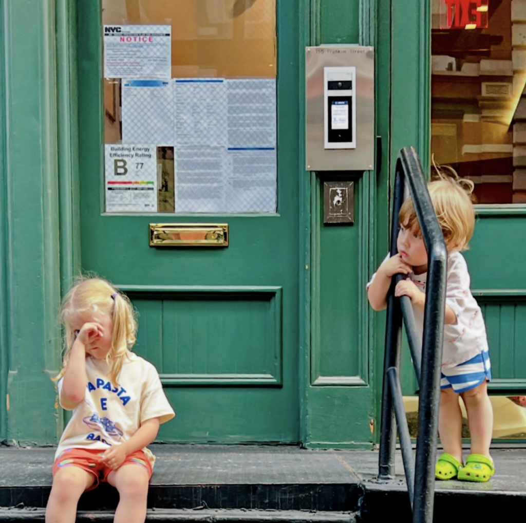 two young children, girl and boy outside a green door, one sitting on steps, the other looking through rail handles