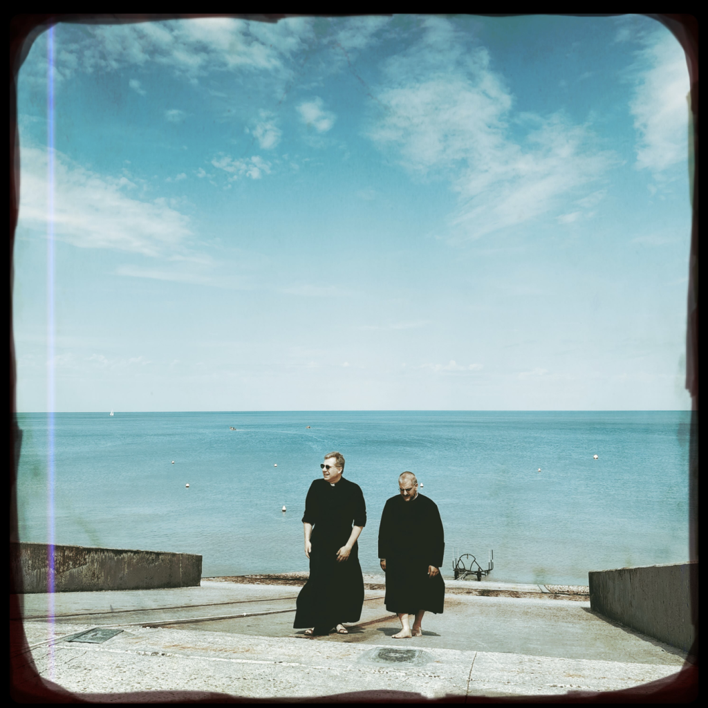 Two priests in black robes walk up a concrete slipway beside a calm turquoise sea beneath a wide blue sky, photographed with a vintage film-style border.