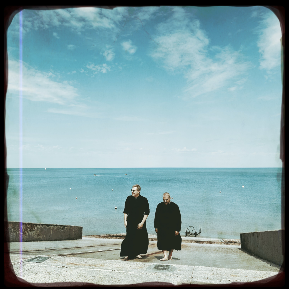 Two priests in black robes walk up a concrete slipway beside a calm turquoise sea beneath a wide blue sky, photographed with a vintage film-style border.