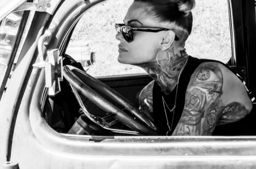 Black-and-white portrait of a tattooed woman wearing sunglasses and hoop earrings, seated behind the wheel of a vintage car and looking through the open window.