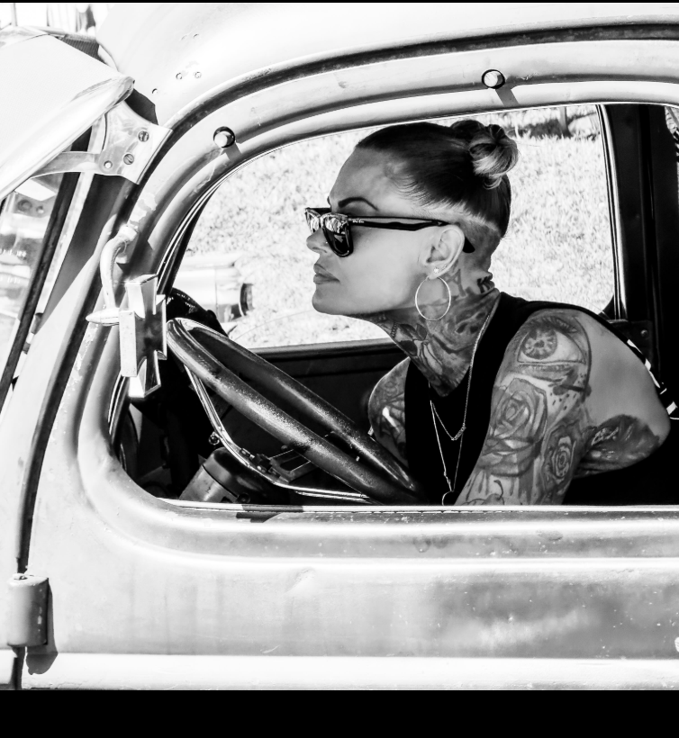 Black-and-white portrait of a tattooed woman wearing sunglasses and hoop earrings, seated behind the wheel of a vintage car and looking through the open window.