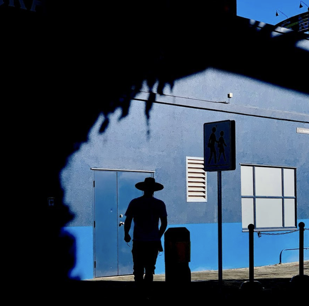 Silhouetted man in a wide-brimmed hat walking past a vivid blue building, framed by deep architectural shadows and strong sunlight.