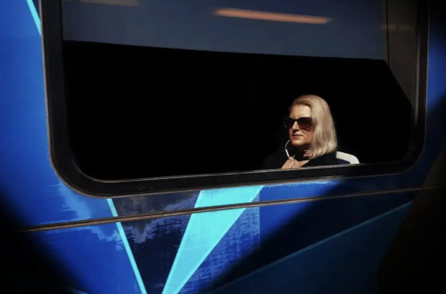 Woman wearing sunglasses seated inside a dark window framed by vivid blue geometric shapes and strong contrasting light.