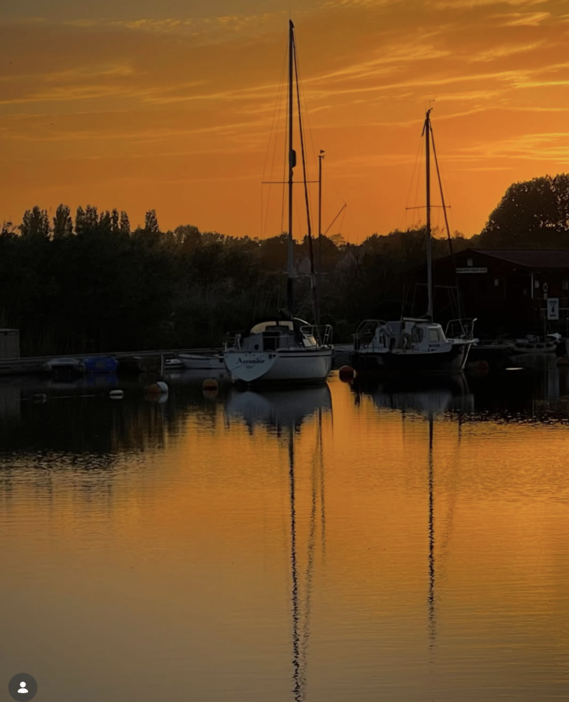 Sailing boats moored on calm water at sunset, silhouetted against a vivid orange sky with their masts reflected in the water.
