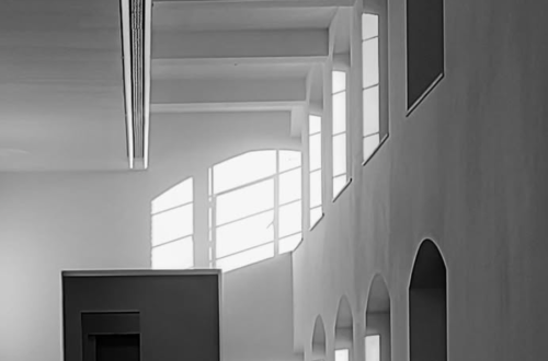 Black-and-white architectural photograph of a lone figure standing beneath tall arched windows, with strong geometric shadows and light falling across a minimalist interior.