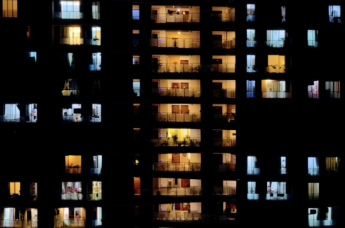 Night photograph of a high-rise apartment building with illuminated windows glowing in warm yellow and cool blue tones against the dark facade.