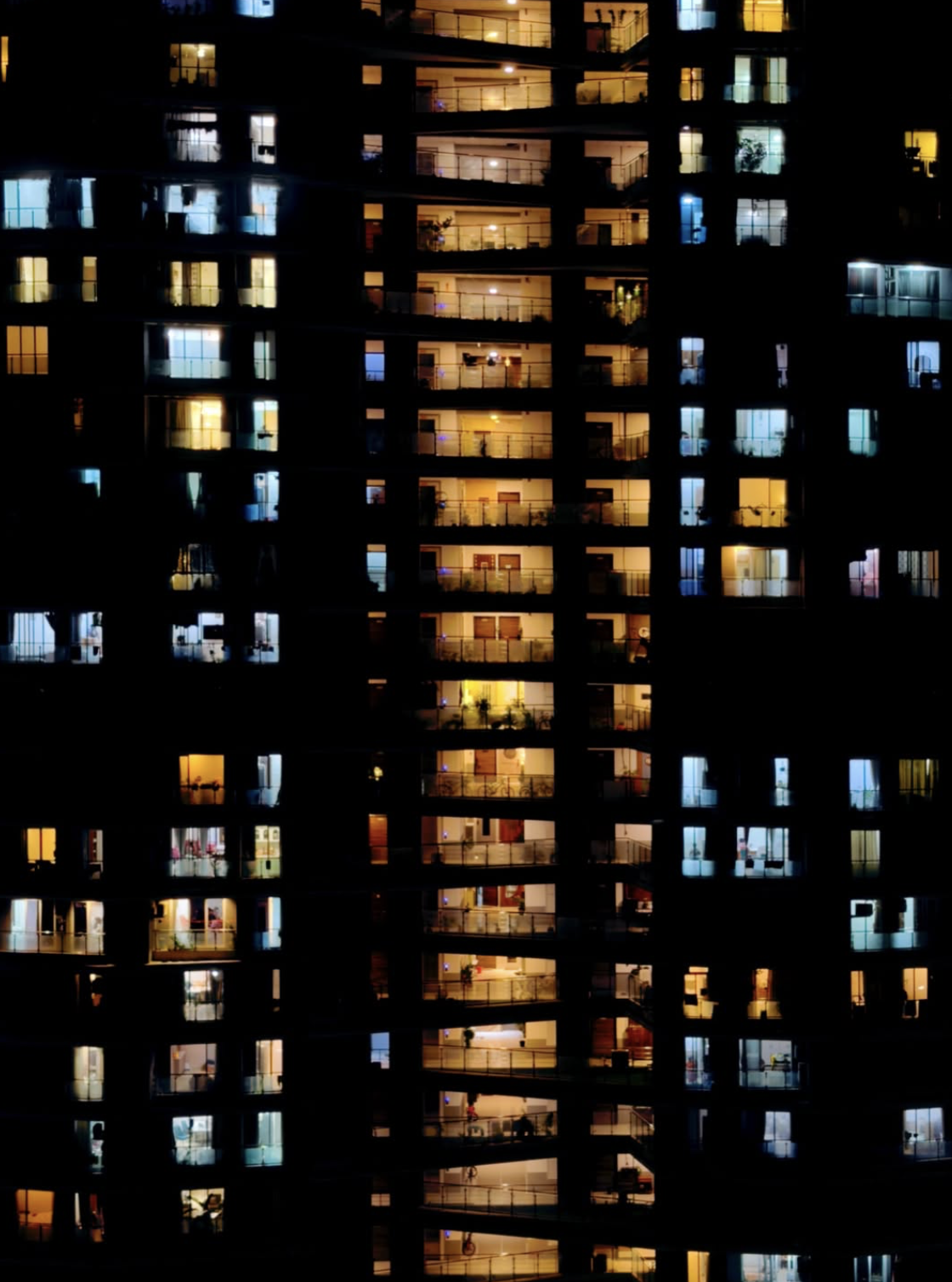 Night photograph of a high-rise apartment building with illuminated windows glowing in warm yellow and cool blue tones against the dark facade.