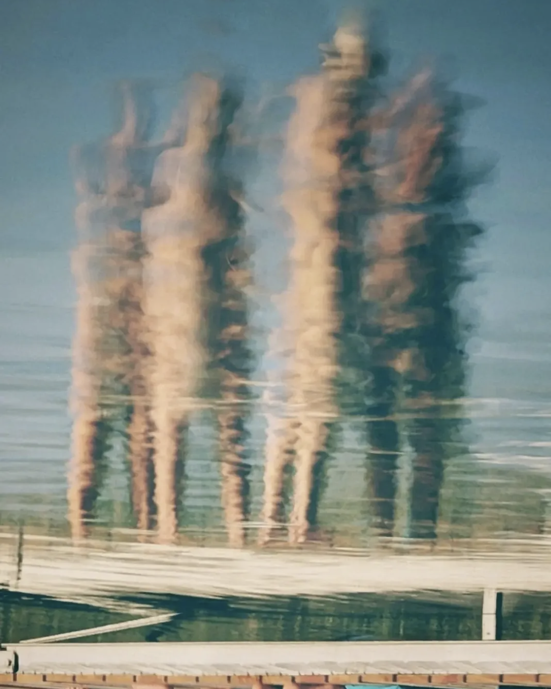 Atmospheric abstract photograph by Carmen Zamos, showing elongated, blurred human figures reflected across a glassy surface in soft blue, teal, peach and cream tones.Atmospheric abstract photograph by Carmen Zamos, showing elongated, blurred human figures reflected across a glassy surface in soft blue, teal, peach and cream tones.