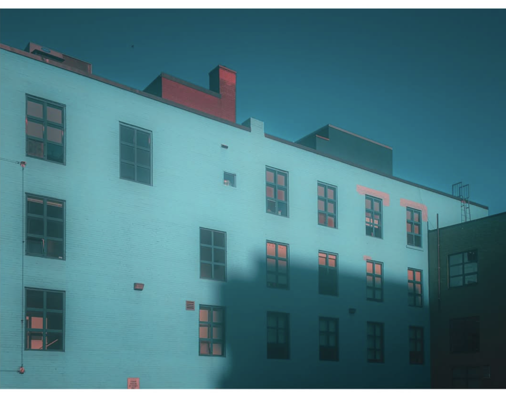 Modern building façade washed in turquoise-blue light, with coral reflections in the windows and a dramatic geometric shadow stretching across the lower floors.