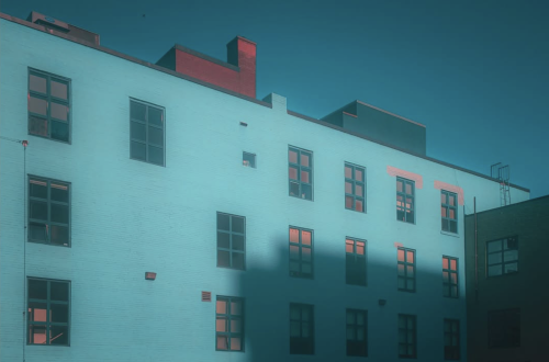Modern building façade washed in turquoise-blue light, with coral reflections in the windows and a dramatic geometric shadow stretching across the lower floors.