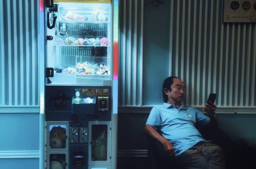 Man seated beside a brightly illuminated Japanese prize machine, looking at his phone in a dark teal interior with striped wall panels.