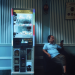 Man seated beside a brightly illuminated Japanese prize machine, looking at his phone in a dark teal interior with striped wall panels.