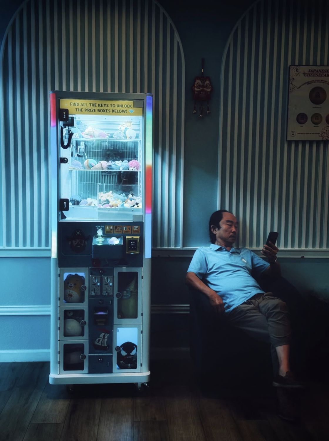 Man seated beside a brightly illuminated Japanese prize machine, looking at his phone in a dark teal interior with striped wall panels.