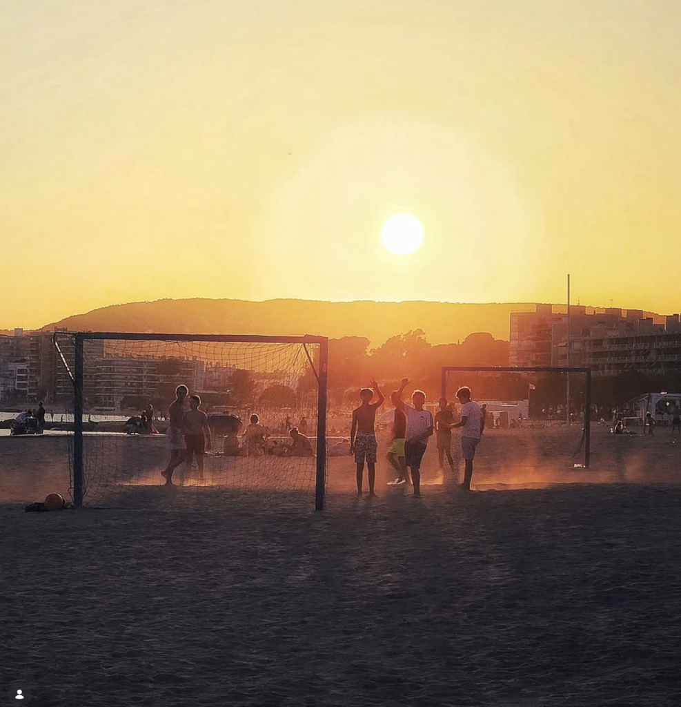 Children playing football on a dusty pitch at sunset, silhouetted between goalposts beneath a glowing golden sky, photographed by s.vizcaino_color for TheAppWhisperer Pic of the Day 2625.