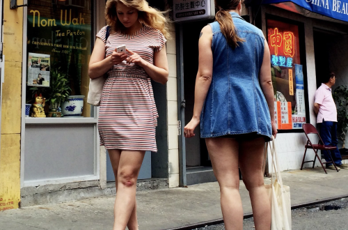 Candid street photograph of two women outside colourful Chinatown storefronts, one looking at her smartphone, surrounded by Chinese signage and neon lights.