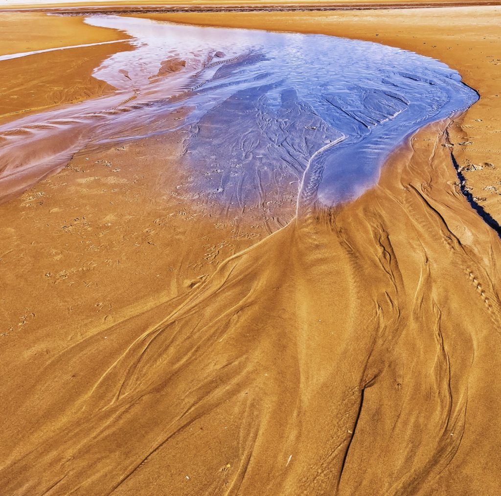 Abstract patterns of blue tidal water flowing across rich golden-orange sand, creating sweeping organic lines and intricate textures across the beach.