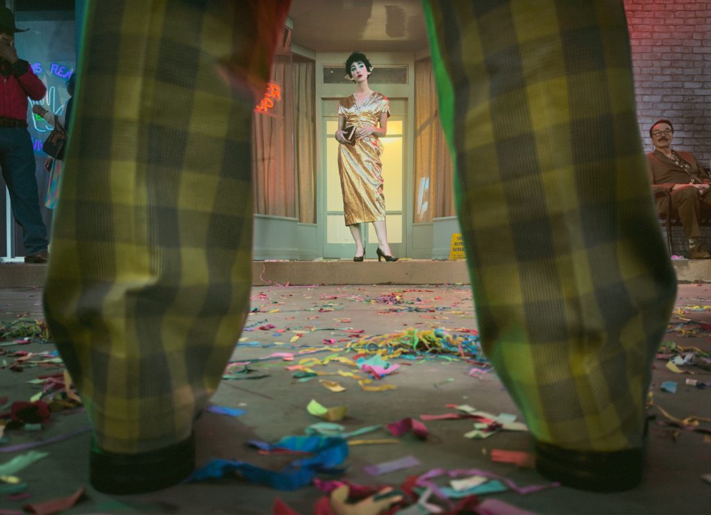 Alex Prager photograph of a woman in a gold dress framed between the legs of a figure in checked trousers, with confetti scattered across the ground.