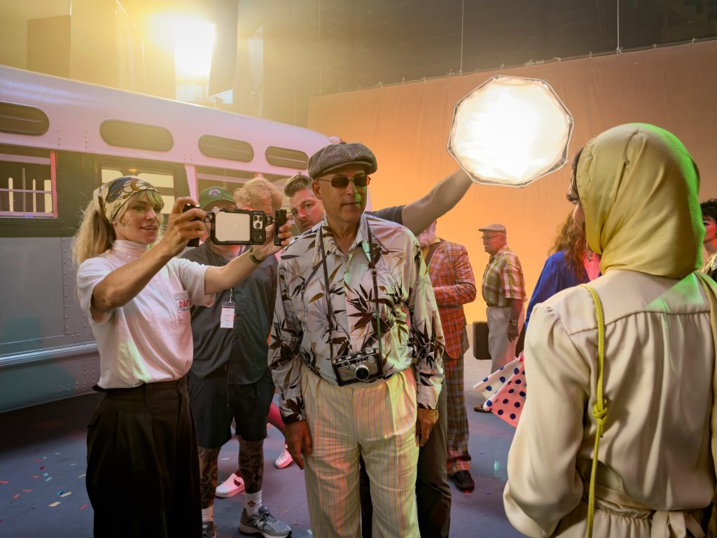 Alex Prager photographing a woman in vintage clothing with an iPhone beside a window illuminated by red neon lettering.