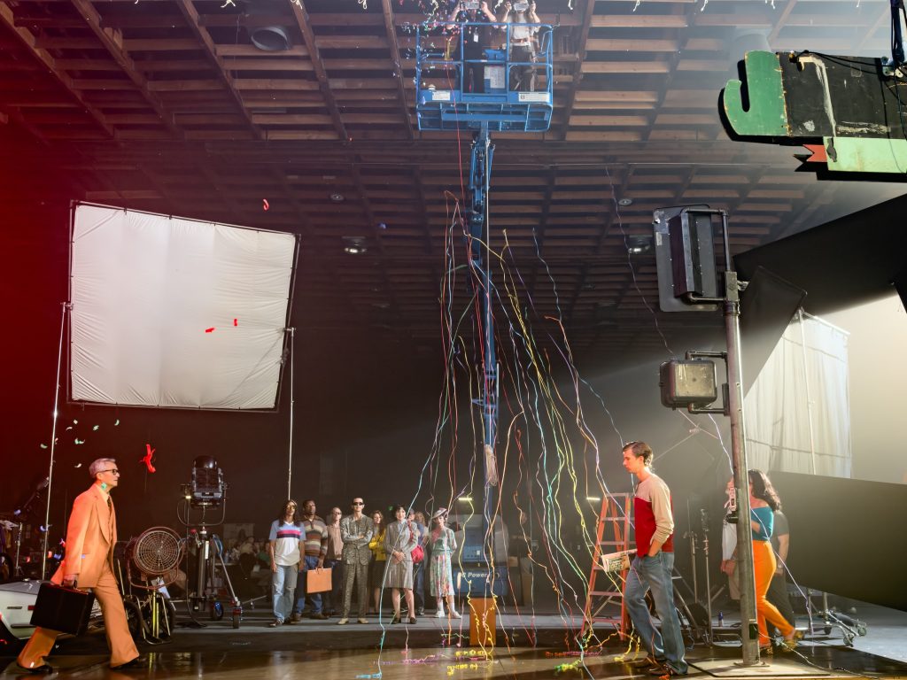 Behind-the-scenes view of Alex Prager photographing a staged street scene as a crew member holds a large studio light.