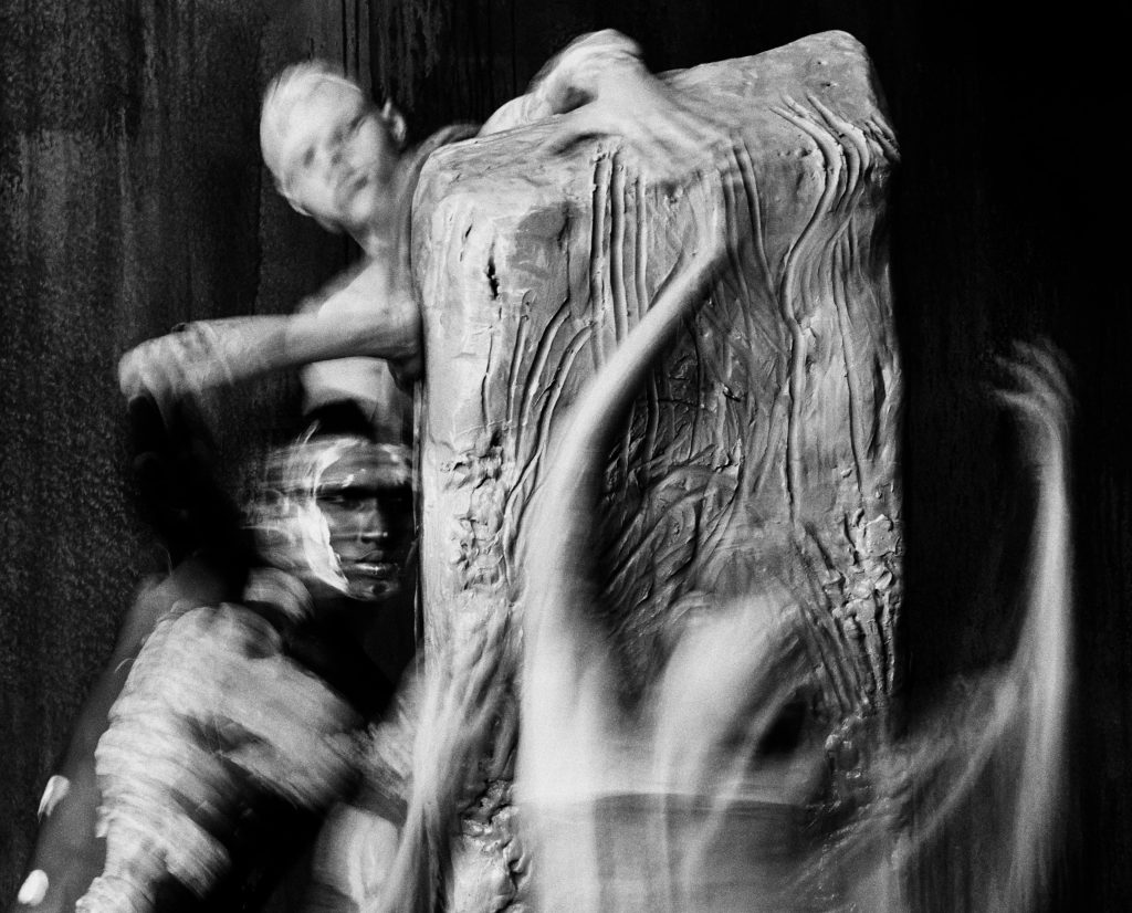 Jack Davison black-and-white photograph of blurred human figures surrounding and reaching across a large textured sculptural form.