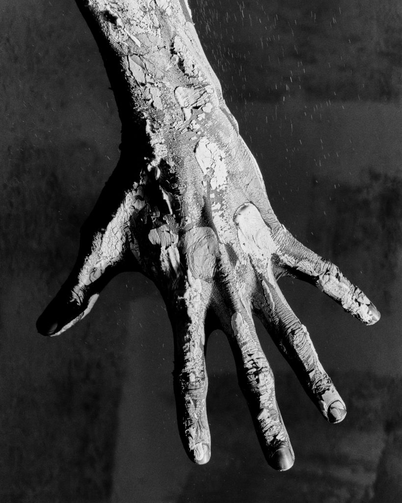 Jack Davison black-and-white close-up of an outstretched hand covered in cracked, peeling material against a dark background.