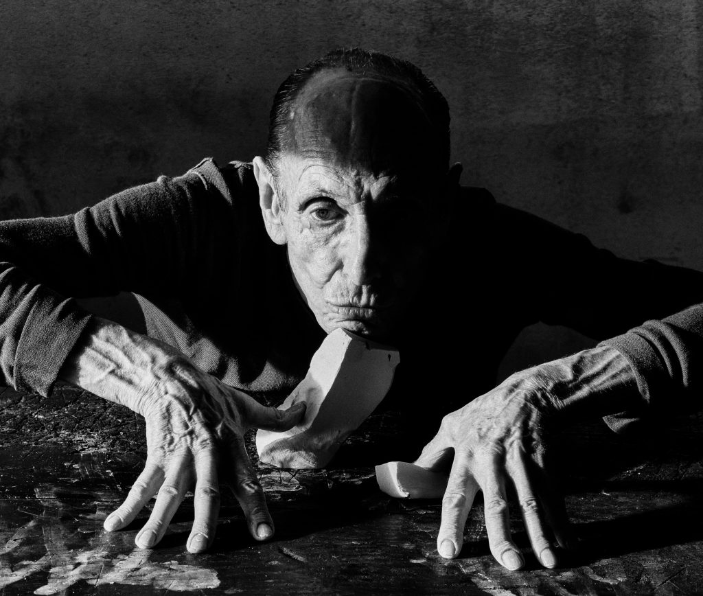 Jack Davison black-and-white portrait of a man leaning across a table with outstretched hands and fragments of a small sculpture before him.