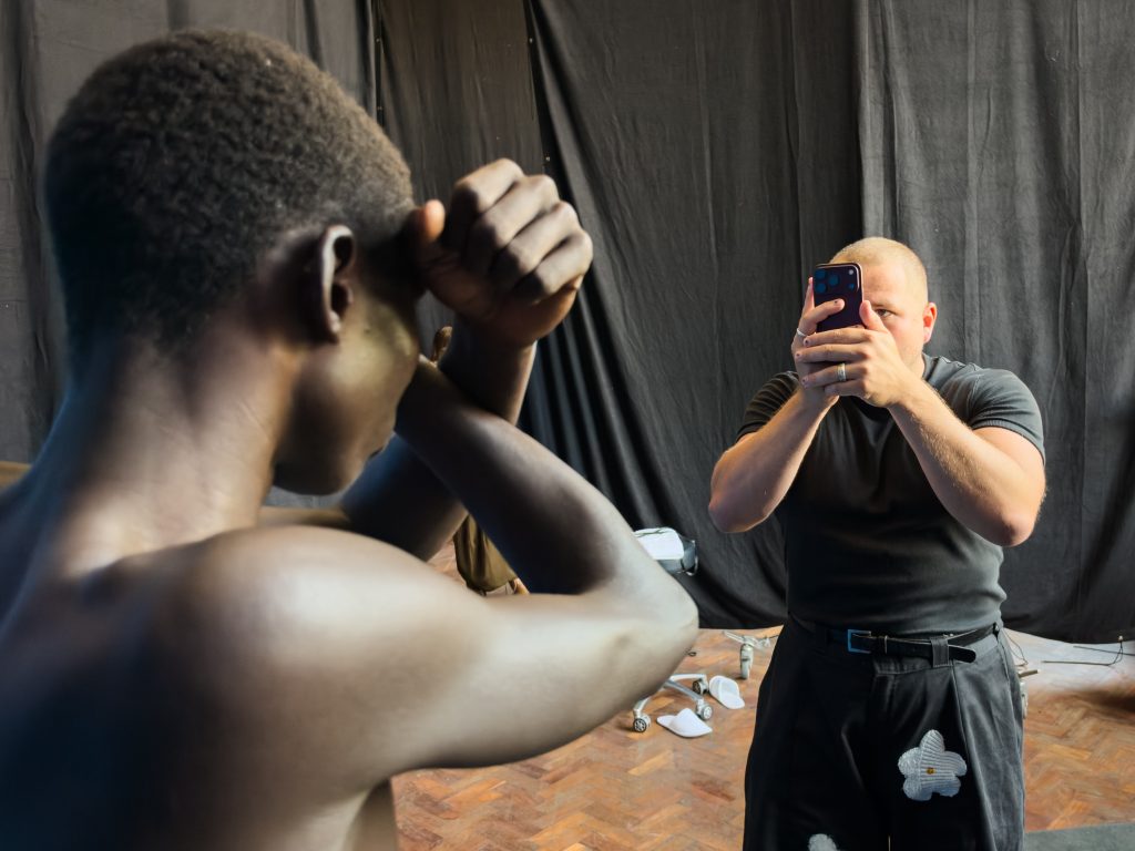 Jack Davison photographing a shirtless model with an iPhone against a black studio backdrop.
