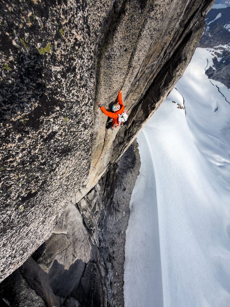 Jimmy Chin photograph looking down on a climber in an orange jacket scaling a steep granite wall high above a snow-covered glacier.