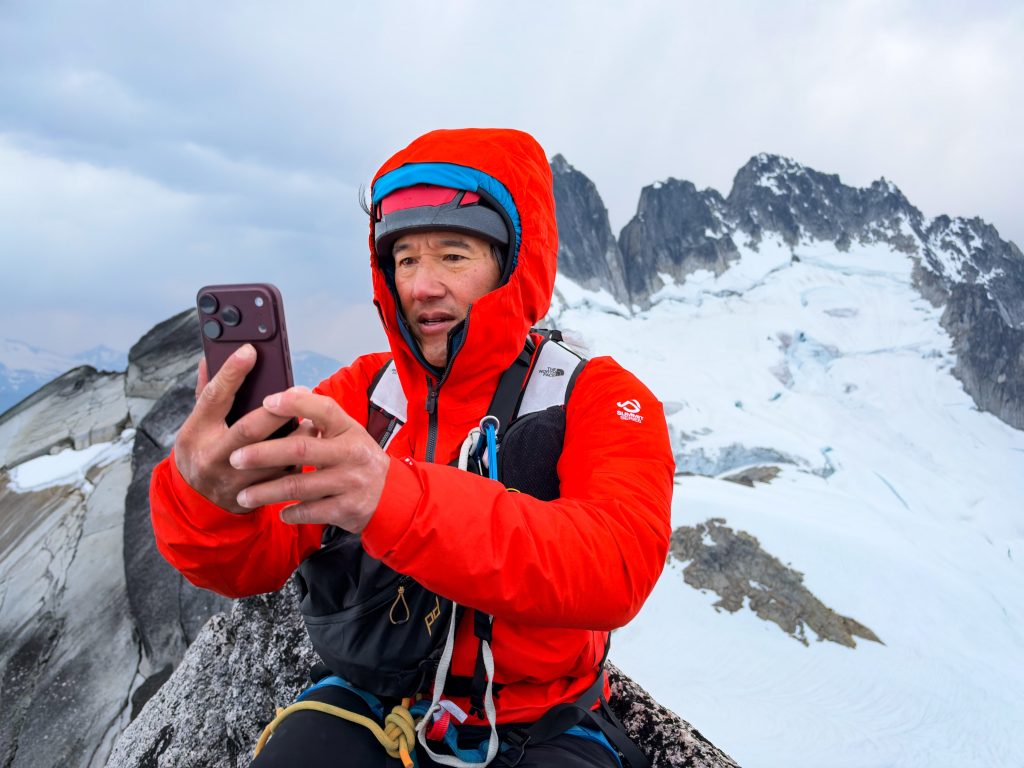 Jimmy Chin photograph of a climber in a bright orange jacket ascending a dramatic granite peak surrounded by snowy mountains beneath a blue, cloud-filled sky.
