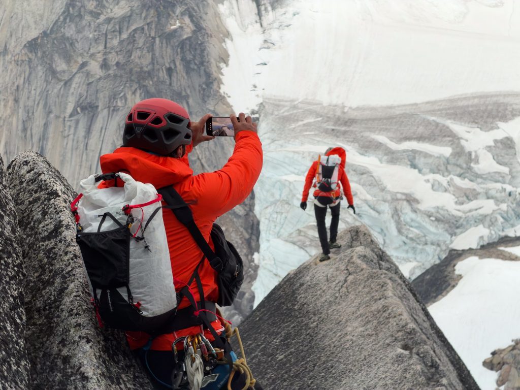 immy Chin photograph looking down on a climber in an orange jacket scaling a steep granite wall high above a snow-covered glacier.