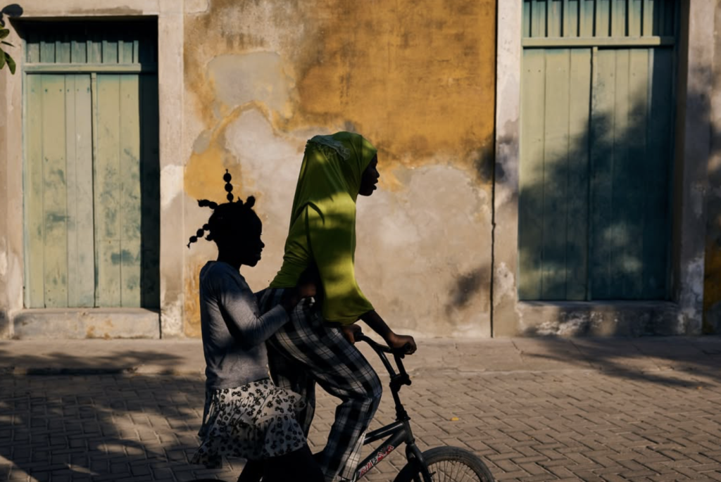Street photograph of two figures travelling together on a bicycle, silhouetted against a sunlit ochre wall with pale green wooden doors. One figure wears a vivid green head covering, while strong afternoon light and deep shadows create a striking graphic composition.