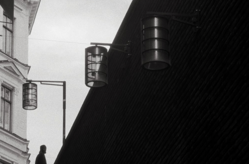 Black-and-white street photograph captured in Stockholm, Sweden, showing a solitary silhouetted figure at the top of a staircase beside a dramatic dark diagonal wall, with hanging cylindrical lights and traditional architecture in the background.