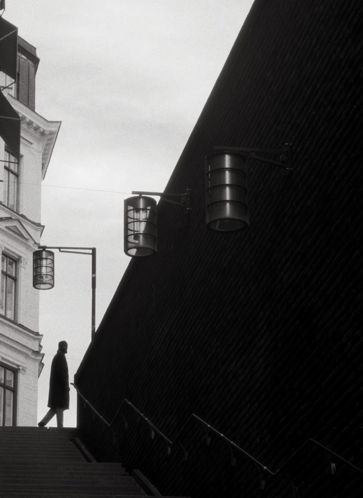 Black-and-white street photograph captured in Stockholm, Sweden, showing a solitary silhouetted figure at the top of a staircase beside a dramatic dark diagonal wall, with hanging cylindrical lights and traditional architecture in the background.