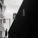 Black-and-white street photograph captured in Stockholm, Sweden, showing a solitary silhouetted figure at the top of a staircase beside a dramatic dark diagonal wall, with hanging cylindrical lights and traditional architecture in the background.