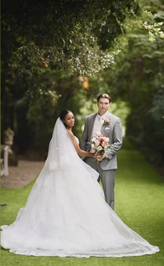 Bride and groom, Jake and Stephanie, posing together in a lush green garden in Italy on their wedding day, with Stephanie wearing a flowing white bridal gown and veil and Jake in a light grey suit holding a bouquet of soft pink and white flowers.