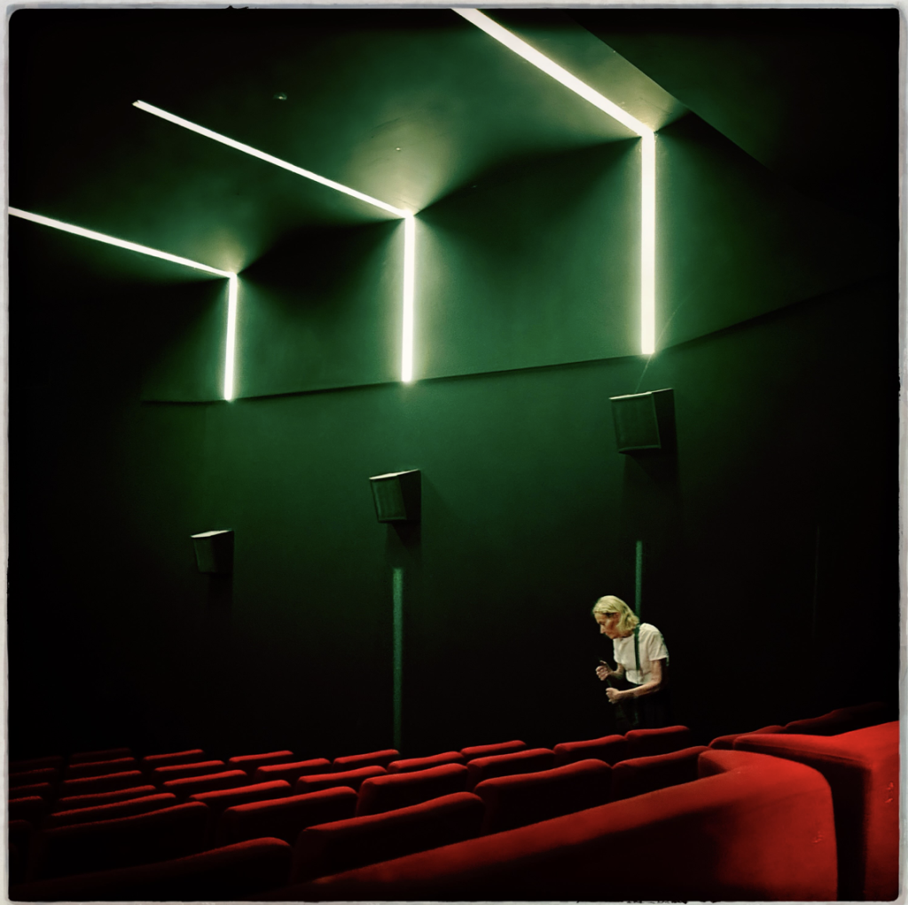 A lone person stands among rows of vivid red cinema seats beneath dramatic geometric green lighting.