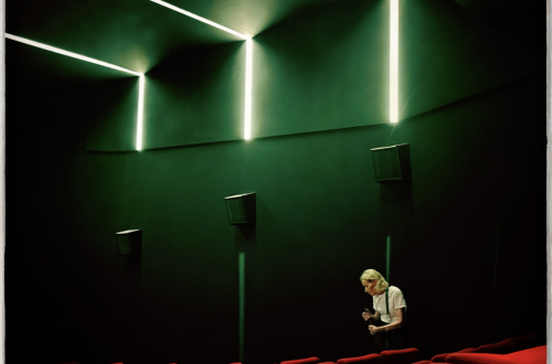 A lone person stands among rows of vivid red cinema seats beneath dramatic geometric green lighting.