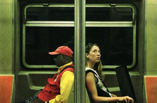 Street photograph inside a New York City subway carriage showing two passengers seated back-to-back on opposite sides of a central metal pole. Strong red, yellow, orange and green tones, geometric seating and the symmetrical composition create a striking visual contrast.