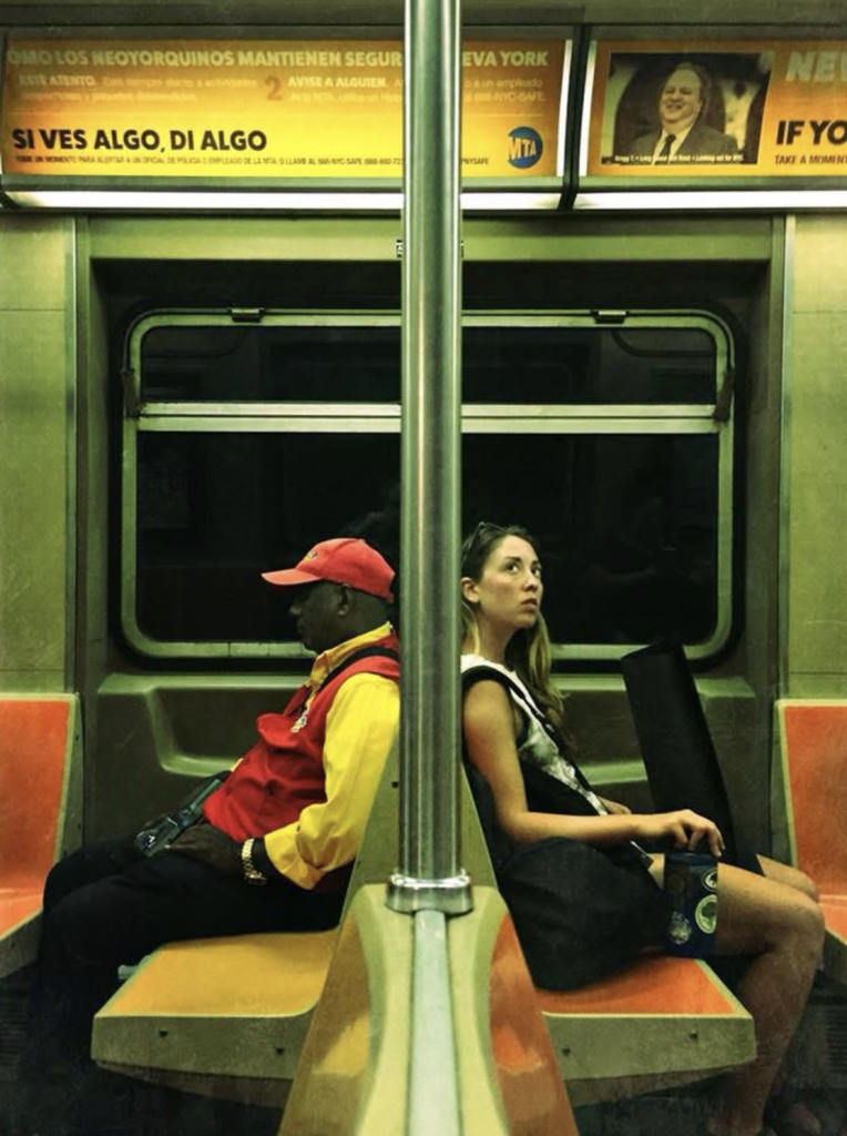 Street photograph inside a New York City subway carriage showing two passengers seated back-to-back on opposite sides of a central metal pole. Strong red, yellow, orange and green tones, geometric seating and the symmetrical composition create a striking visual contrast.