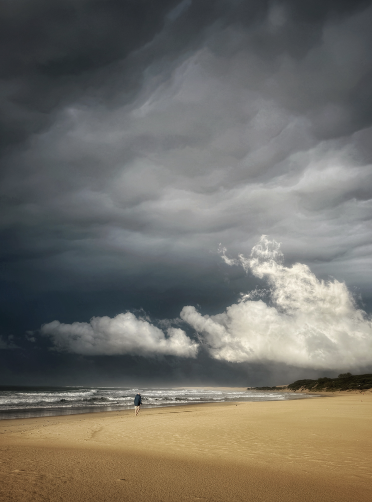 A lone figure walks along a wide golden beach beneath towering dark storm clouds, with white waves breaking along the shoreline.